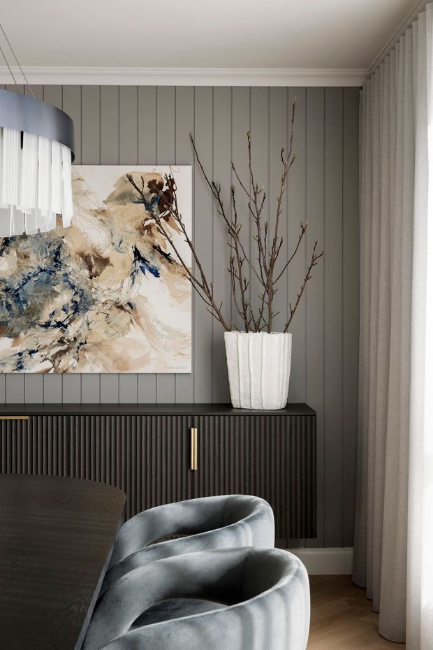 A refined dining or living area featuring a dark grey vertical-panelled feature wall, a fluted dark built-in sideboard with brass handles, abstract artwork, sculptural grey upholstered chairs, and a dark timber table. Floor-to-ceiling curtains and a sculptural pendant contribute to the softly lit, sophisticated interior.