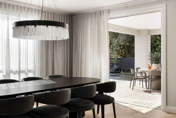 Contemporary dining room with a large dark oval dining table and upholstered black dining chairs, illuminated by a sculptural circular pendant light. Floor-to-ceiling sheer and grey curtains frame a wide opening to a sunlit outdoor dining terrace, with neutral walls, pale timber flooring, and understated built-in detailing.