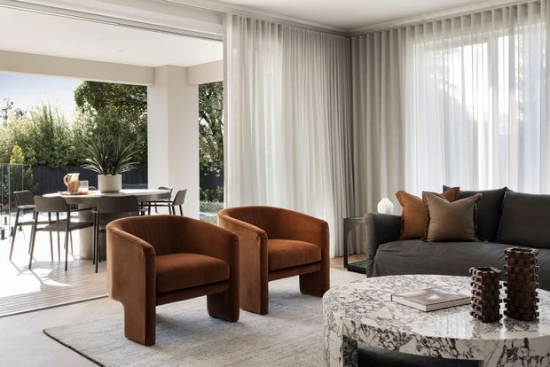 A bright, refined open-plan living and dining area with floor-to-ceiling sheer curtains, large windows, a dark sectional sofa, two rust-brown upholstered armchairs, and a round marble coffee table. A dining table and chairs are visible beyond the living area, with greenery outside.