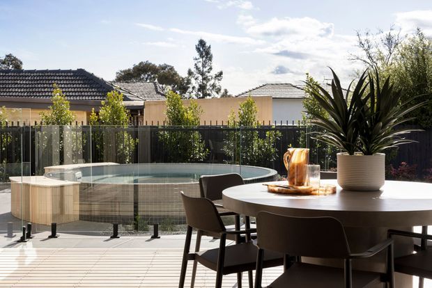 A contemporary outdoor dining area on a timber deck, with a large round dining table and dark chairs in the foreground. Beyond the black-framed privacy fencing is a curved tiled swimming pool with a glass balustrade and a raised spa or seating platform. Low residential buildings with tiled roofs and landscaped planting form the background.
