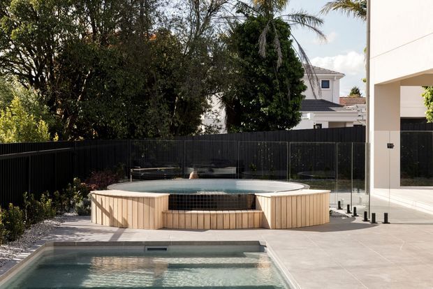 Contemporary residential outdoor area featuring a raised circular spa or plunge pool clad in vertical light timber, a larger rectangular pool in the foreground, pale tiled paving, frameless glass safety fencing, dark boundary fencing, and dense established planting. A modern light-coloured house is partially visible at right and in the background.