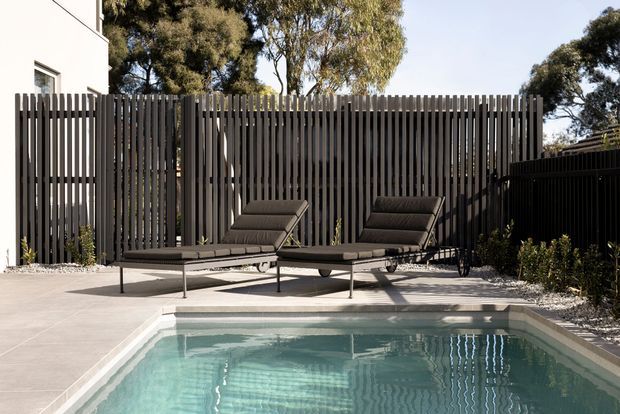 A modern outdoor pool area with a rectangular swimming pool, pale paved surrounds, two dark reclining loungers, and a tall vertical-slat privacy fence. Mature trees and planting are visible beyond the fence, with part of a light-coloured building at left.