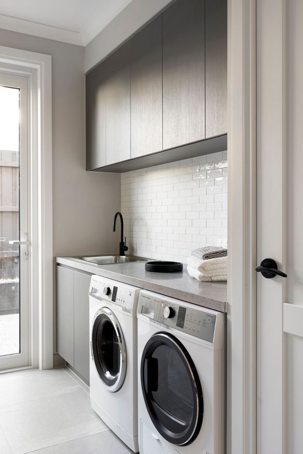 A compact, contemporary laundry room viewed through an open doorway. It features stacked-height dark grey overhead cabinetry, pale grey lower cabinetry, a grey benchtop with integrated stainless sink and black gooseneck tap, white subway-tile splashback, and side-by-side white front-loading washing machine and dryer. A glazed exterior door provides natural light, while folded towels sit on the benchtop.
