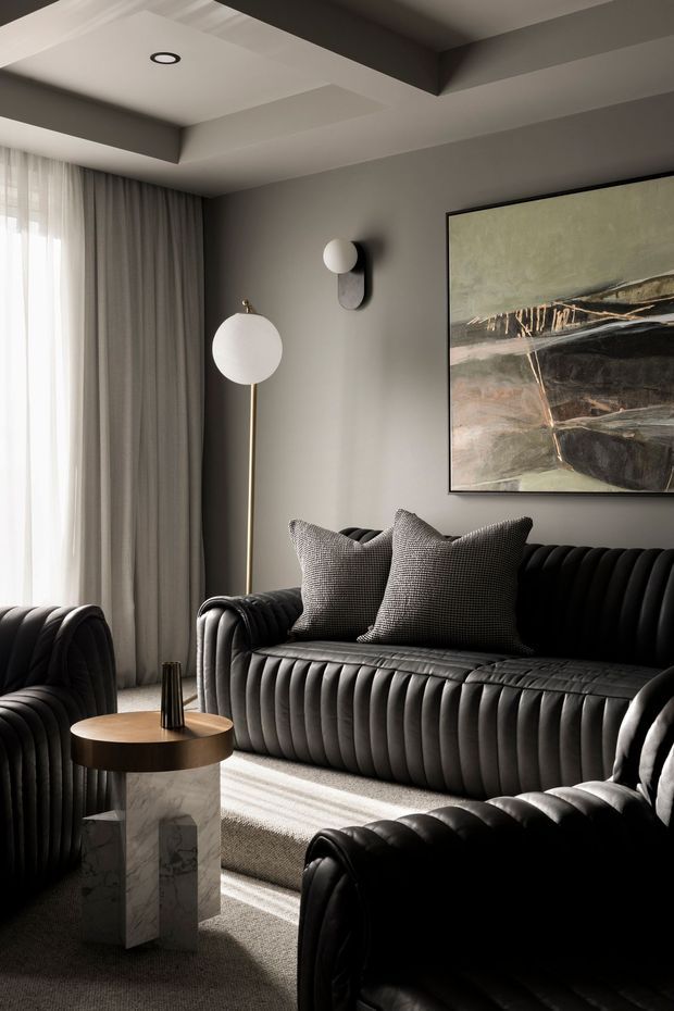 A refined living room with a dramatic black channel-tufted sofa and matching armchairs, grey walls, layered sheer and blackout curtains, sculptural wall and floor lamps, abstract artwork, and a small round wood-and-marble side table. Strong sunlight creates high-contrast shadows across the neutral carpet and dark upholstery. The ceiling features recessed lighting and substantial boxed beams.