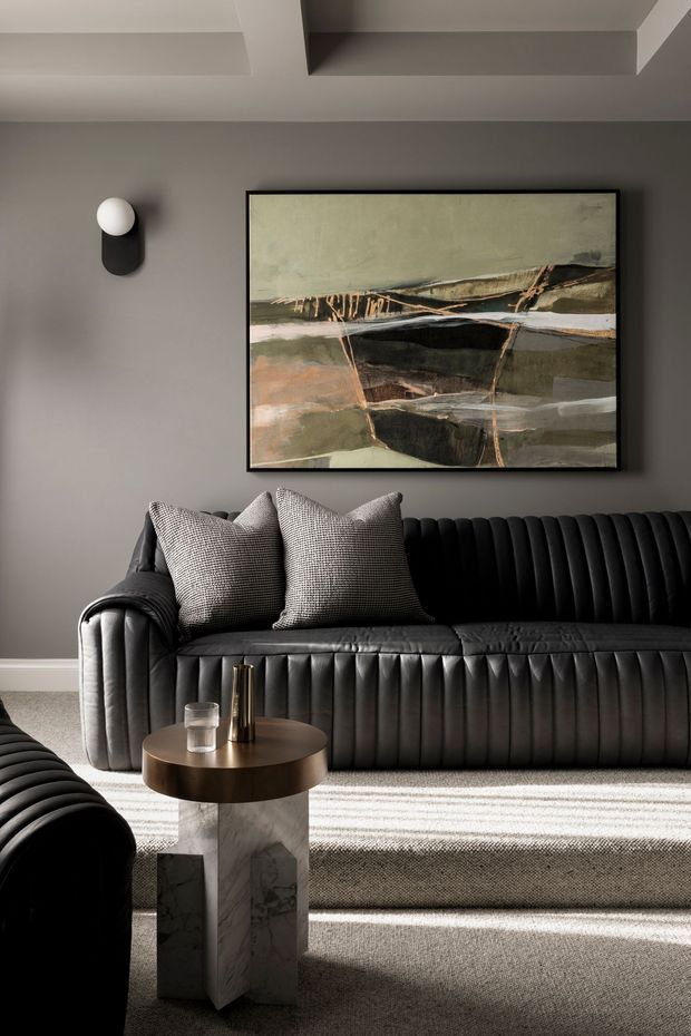 A sophisticated living room with a dark grey wall, sculptural abstract artwork, black vertically channelled leather sofa, matching seating in the foreground, textured neutral rug, and a round bronze-toned coffee table on a marble pedestal base. A modern wall sconce and geometric ceiling detail complete the refined, moody interior.