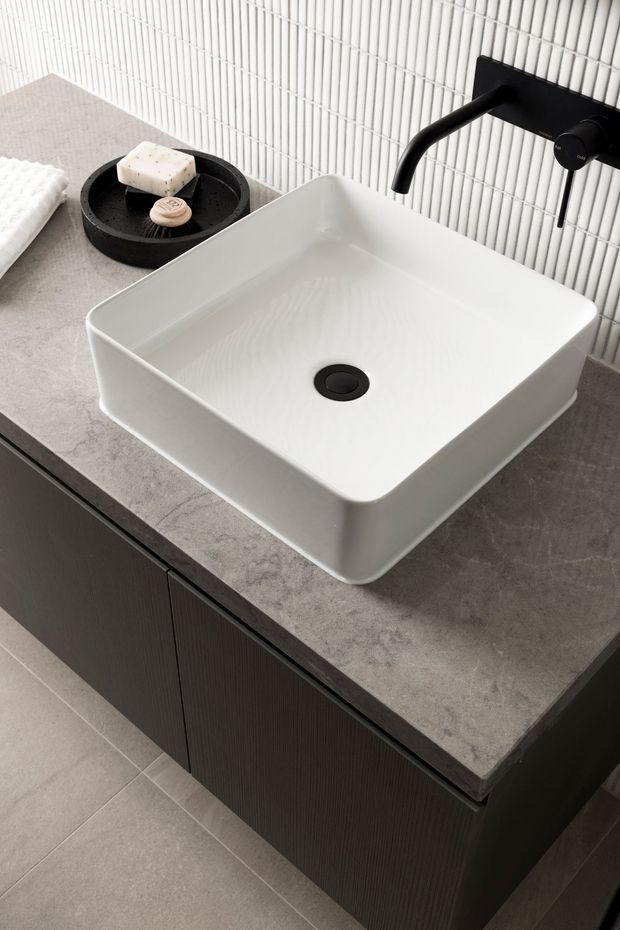 Contemporary bathroom vanity featuring a large white vessel basin, matte black wall-mounted tapware, dark vertically textured cabinetry, a grey stone-look benchtop, and white vertically fluted wall tiles. The composition is a close-up product-style view of the vanity area.