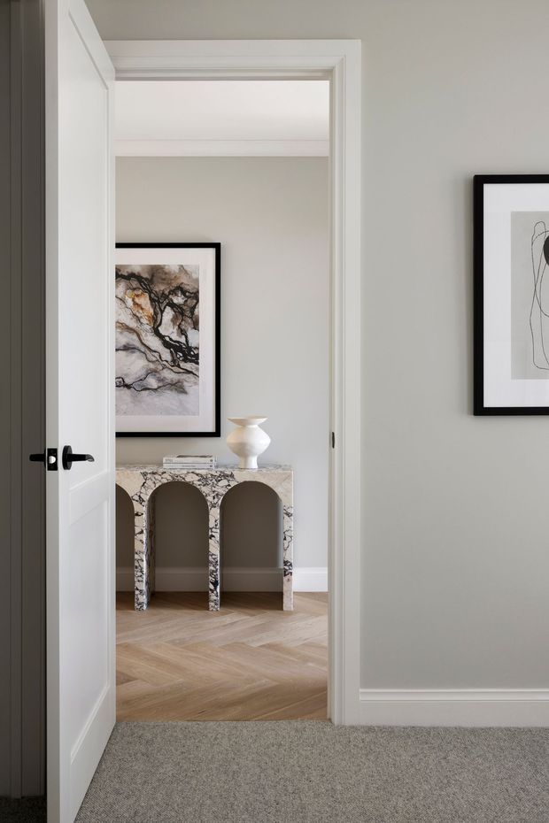 View through an open white paneled door into a refined interior, featuring a marble console with arched openings, framed abstract artwork, a sculptural white vase, light herringbone timber flooring, pale grey walls, and grey carpet in the foreground. The space has crisp white trim and black accents.