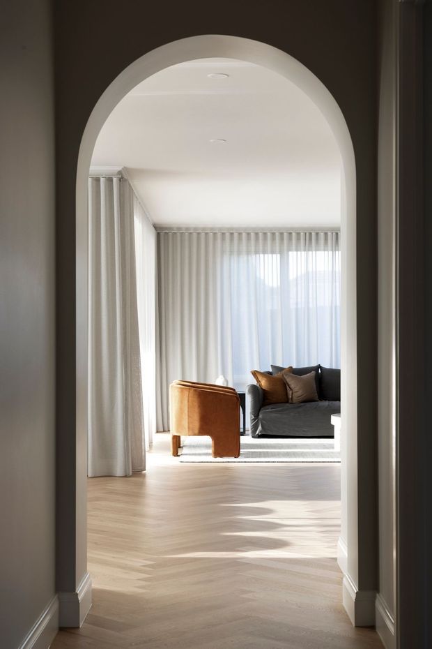 A bright, refined living room viewed through a tall rounded archway. The room features pale herringbone timber flooring, full-height sheer curtains, a dark charcoal sofa with warm brown and neutral cushions, and a caramel leather lounge chair. Soft daylight creates a calm, elegant atmosphere, with subtle traditional architectural detailing around the arch and skirting.