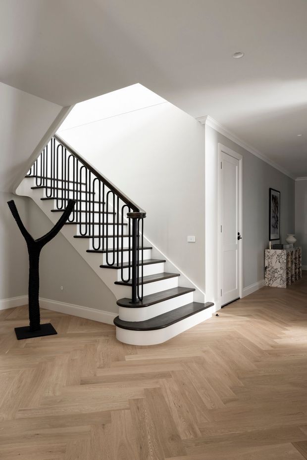 Bright, refined residential hallway featuring a sculptural staircase with dark timber treads, curved white risers and black metal balustrades. The space has pale herringbone timber flooring, soft grey walls, white trim and doors, recessed ceiling lighting, and a decorative console with artwork along the corridor.