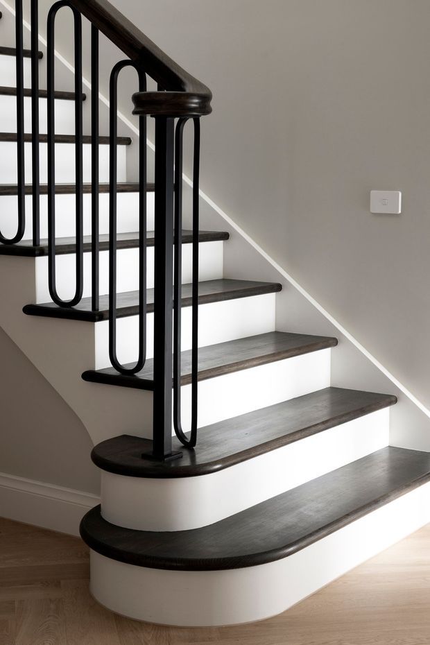 A refined staircase with dark timber treads and handrail, white curved stair risers and landings, and distinctive black metal looping balustrade. The staircase sits against a light grey wall with pale timber flooring, creating a clean, high-contrast contemporary interior.