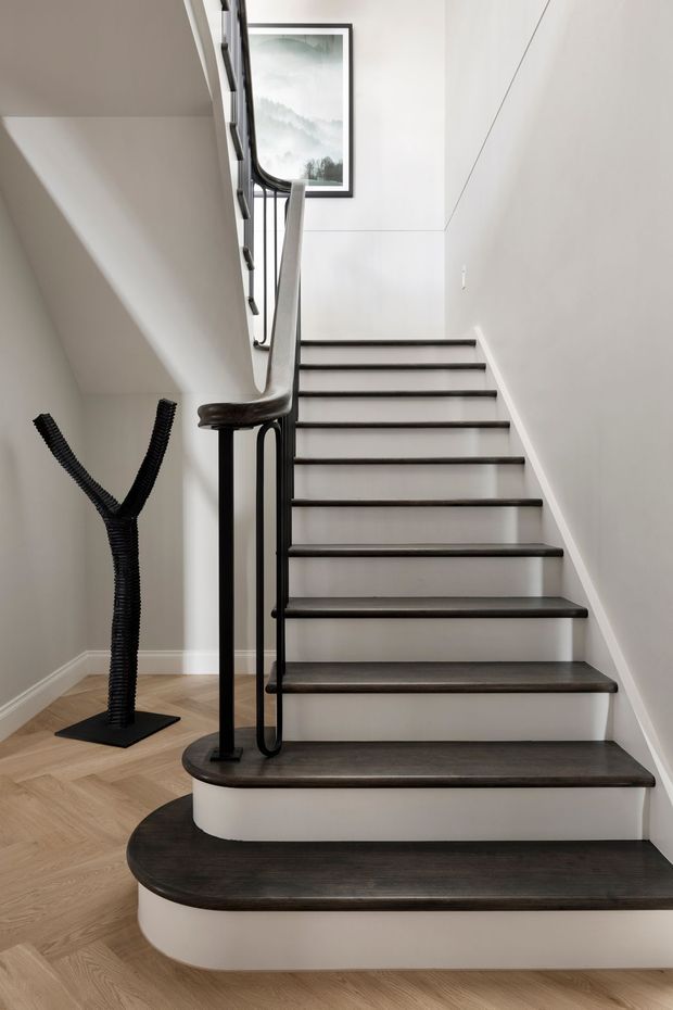 A refined interior stairway with dark timber treads and rounded stair edges, white painted walls and risers, and a distinctive black metal handrail and balustrade. The stair curves around a central landing, with light herringbone timber flooring and a minimalist black sculptural object beneath the stairs. A framed artwork is visible on the upper wall.