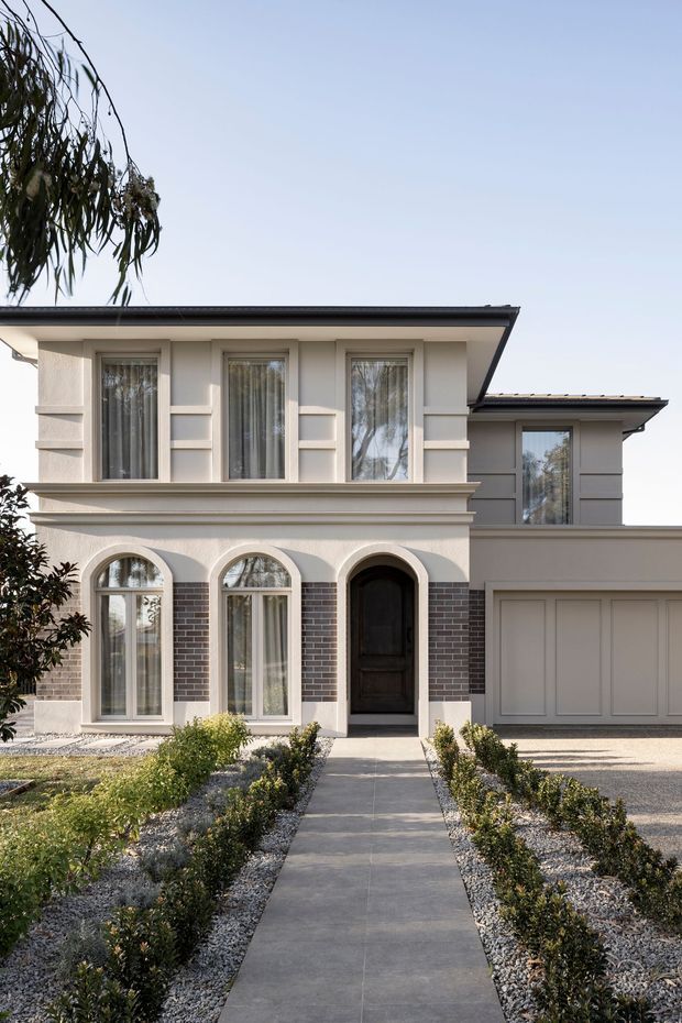 Front exterior view of a refined two-storey residence with a pale rendered facade, dark low-pitched roof, symmetrical upper-storey windows, arched ground-floor windows and entry, subtle brick surrounds, and an integrated garage. A formal landscaped front garden with clipped hedges and a central paved path leads to the entrance.