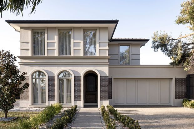 Exterior view of a two-storey residence with a symmetrical rendered facade, dark brick accents, arched ground-floor windows, tall upper-level windows, a recessed dark entry door, and an integrated double garage. The home has a low-pitched roof with broad eaves and a formal landscaped front approach.