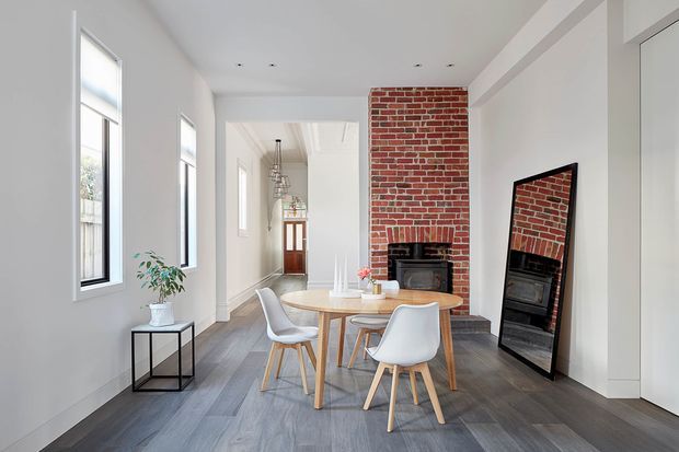 Bright contemporary dining area with a round timber dining table and white upholstered chairs, arranged in front of an exposed red-brick fireplace. Tall windows, pale walls, grey timber-look flooring, and a large leaning mirror create an airy, refined interior, with a hallway and entry door visible beyond.