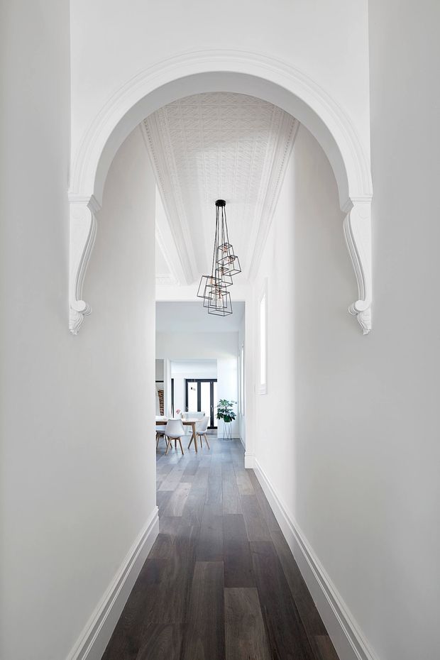 A bright, narrow residential hallway framed by an ornate white arched opening with decorative plaster moulding and a patterned pressed-tin or plaster ceiling. Dark timber flooring leads toward a light-filled dining area with a small round table, chairs, pendant light, and a potted plant. The space combines heritage detailing with a clean contemporary interior.
