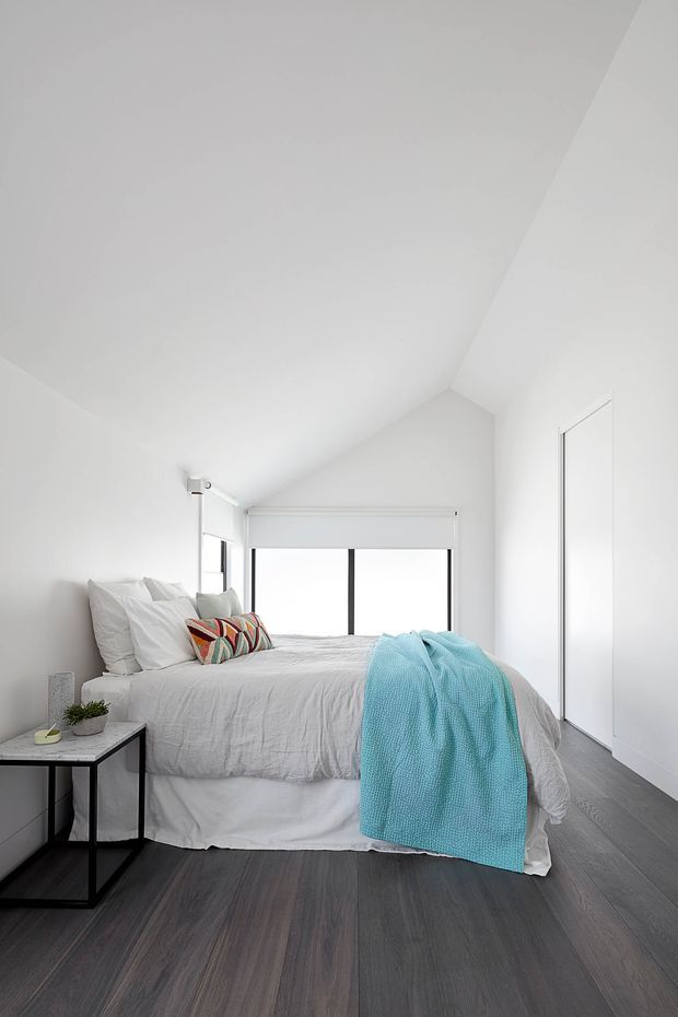 Bright minimalist bedroom with a pitched, vaulted ceiling, white walls, and a large rear window. The room features a neatly made bed with white bedding, a colorful patterned accent cushion, and a turquoise throw. Dark timber-look flooring, a slim black-framed bedside table, and concealed white built-in wardrobe doors complete the clean contemporary interior.