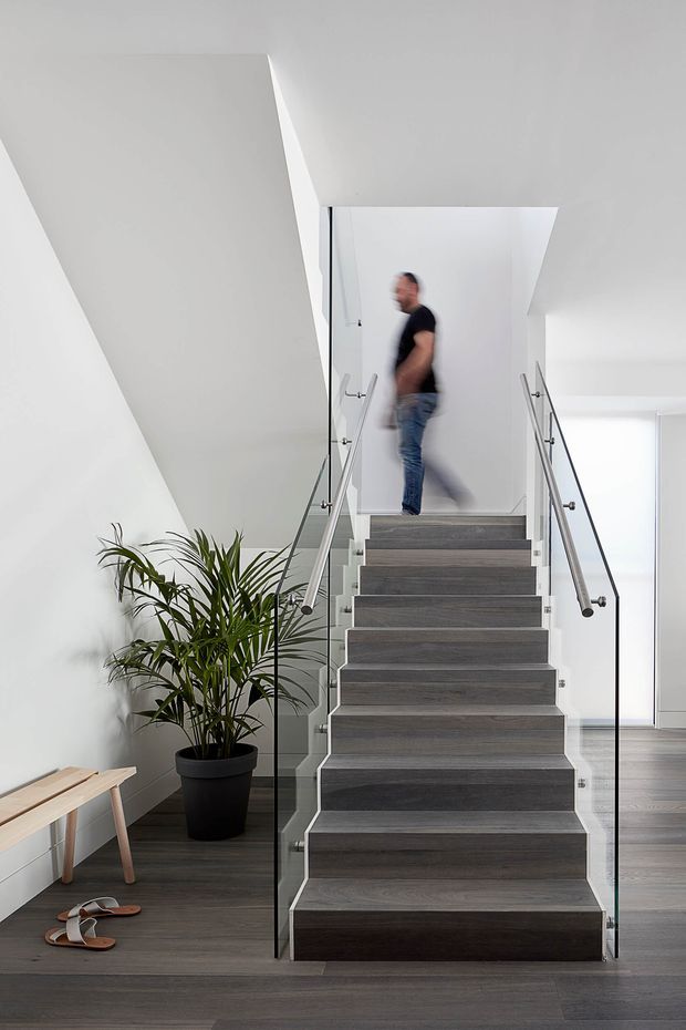 Bright contemporary interior centered on a straight staircase with dark grey timber-look treads and matching flooring. Clear glass balustrades with slim metal handrails flank the stairs, while white angular walls and ceiling planes create a clean architectural composition. A potted palm and small bench occupy the lower landing, and a blurred person is visible on the upper landing.