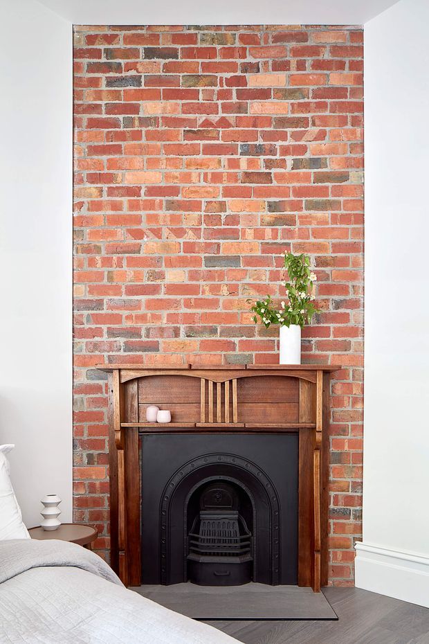 A bright bedroom featuring a restored exposed red-brick chimney breast and traditional timber fireplace surround with a black cast-iron insert. The fireplace is styled with a small white vase of greenery and simple decor, while part of a bed and bedside table are visible in the foreground. White walls, white skirting boards, and dark grey flooring create a clean contrast with the warm brick and timber.