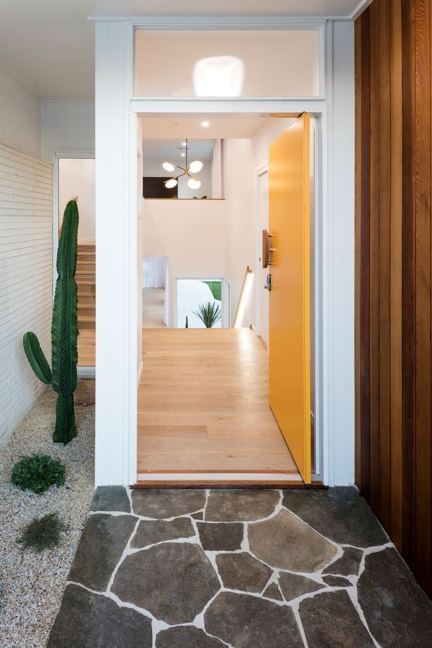 View from outside toward a residential entryway with a bright yellow open front door, white painted trim and transom, warm timber flooring, and a distinctive irregular natural-stone threshold or porch. A gravel-and-cactus landscape is visible to the left, while dark vertical timber cladding frames the right side. The interior beyond includes a stairway and contemporary pendant light.