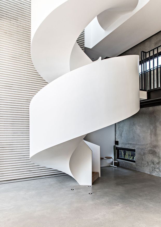 A dramatic interior view dominated by a sculptural white spiral staircase with broad curved balustrades and treads. The staircase rises through a tall void beside an upper-level black metal railing. The space has a polished or smooth grey concrete floor, a textured grey concrete wall, a tall ribbed translucent or glazed wall, and strong natural light from above. The composition emphasizes sweeping curves, double-height volume, and minimalist industrial detailing.