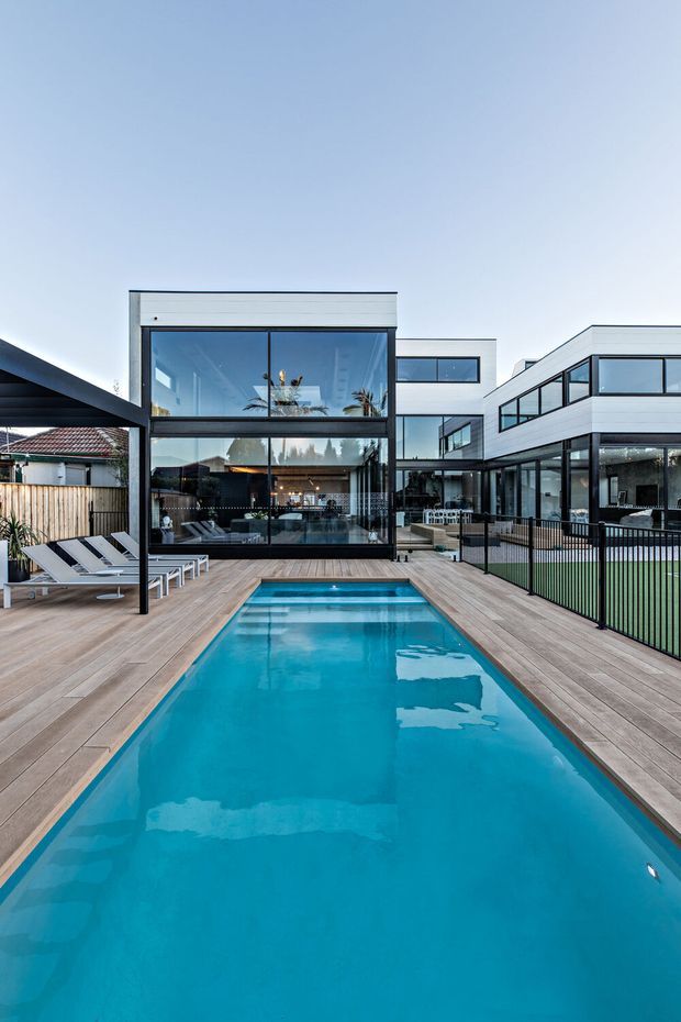 Contemporary multi-level residential property viewed from a pool courtyard. A long turquoise swimming pool is surrounded by a timber deck, with white sun loungers at left and dark metal fencing at right. The building features extensive floor-to-ceiling glazing, white horizontal cladding, dark window frames, flat rooflines, and open-plan interiors visible through the glass. A neighbouring tiled-roof house is visible beyond the fence.