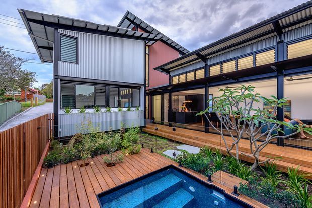 Exterior view of a contemporary multi-level residence arranged around a timber deck and compact swimming pool. The building combines corrugated metal cladding, dark structural framing, large glazed openings, louvred screens and warm timber decking, with planted garden beds integrated around the pool.