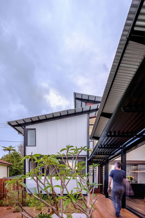 Exterior architectural view of a contemporary multi-level home arranged around a landscaped courtyard. The building features corrugated metal wall cladding, dark metal framing, broad roof overhangs and a covered timber deck or walkway with a slatted pergola. Tropical planting and timber decking soften the modern industrial forms, while a person is visible walking through the courtyard.