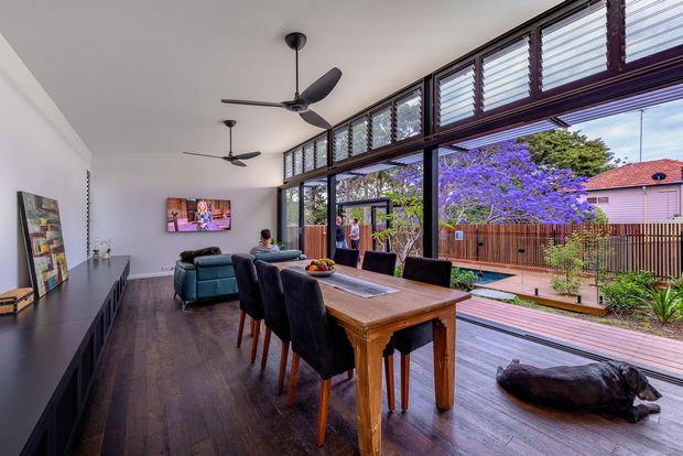 A bright, open-plan dining and living area with a long timber dining table, upholstered dining chairs, teal sofa, dark built-in media cabinetry, ceiling fans, and full-height glazing with high-level louvres. The glazing opens toward a timber deck and landscaped courtyard, with a vivid purple flowering tree visible outside. Dark timber flooring and black-framed glazing create a contemporary indoor-outdoor connection.