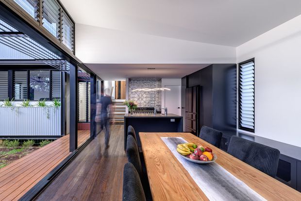 Contemporary open-plan dining and kitchen interior with a long solid timber dining table, dark upholstered dining chairs, dark kitchen cabinetry, and a textured stone-look splashback. Full-height black-framed glazing opens toward a timber deck and planted courtyard, while louvred windows provide filtered light and ventilation. The room has a refined, warm modern character with strong indoor-outdoor connection.