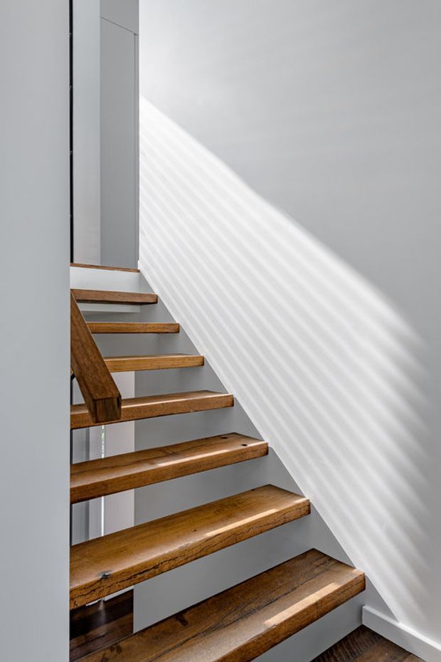 A minimalist interior stairway with floating, open-riser timber treads and a matching timber handrail against smooth white walls. Strong diagonal sunlight creates striped shadows across the wall, emphasizing the clean geometric form and vertical circulation space.