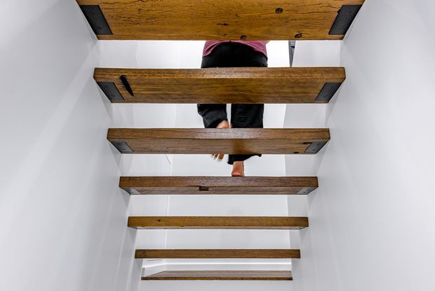 View looking upward through an open-riser staircase with thick exposed timber treads, dark metal side brackets, and crisp white walls. A person is partially visible ascending the stairs. The composition emphasises the stair structure and the contrast between warm wood and white surfaces.