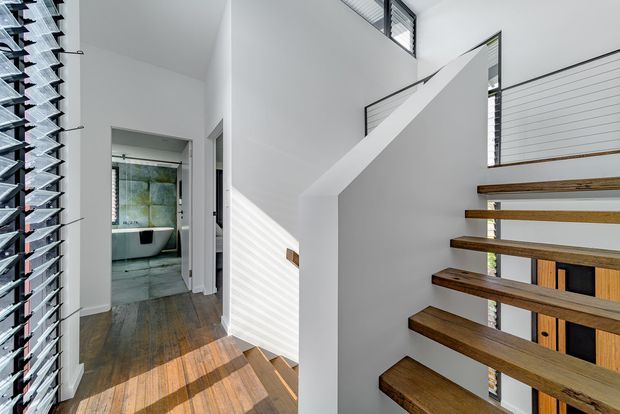 A bright contemporary stairway and hallway with floating dark timber stair treads, white walls, and strong sunlight filtered through louvred windows. A bathroom is visible through the hallway opening, while the stair void includes slim horizontal window openings and a simple white balustrade.