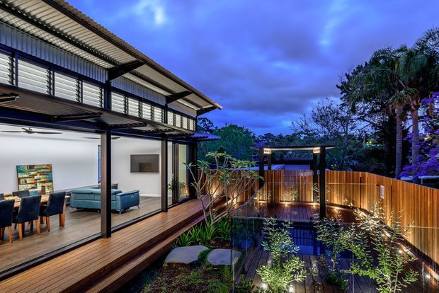 Twilight exterior view of a contemporary multi-level home opening onto a timber deck and landscaped courtyard. The facade features extensive glazing, dark slender framing, horizontal louvres, and broad corrugated roof overhangs. Warm interior and garden lighting illuminate the timber decking, planted courtyard, privacy fencing, and a small covered outdoor structure.
