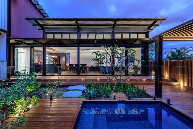 Twilight view of a contemporary multi-level home centred on a landscaped courtyard and reflecting pool. The facade combines dark framed glazing, corrugated metal wall cladding, timber decking, a broad slatted metal canopy, and a covered outdoor entertaining area. Interior living and dining spaces are visible through the glazing, while layered garden planting, stepping stones, and integrated lighting surround the pool.