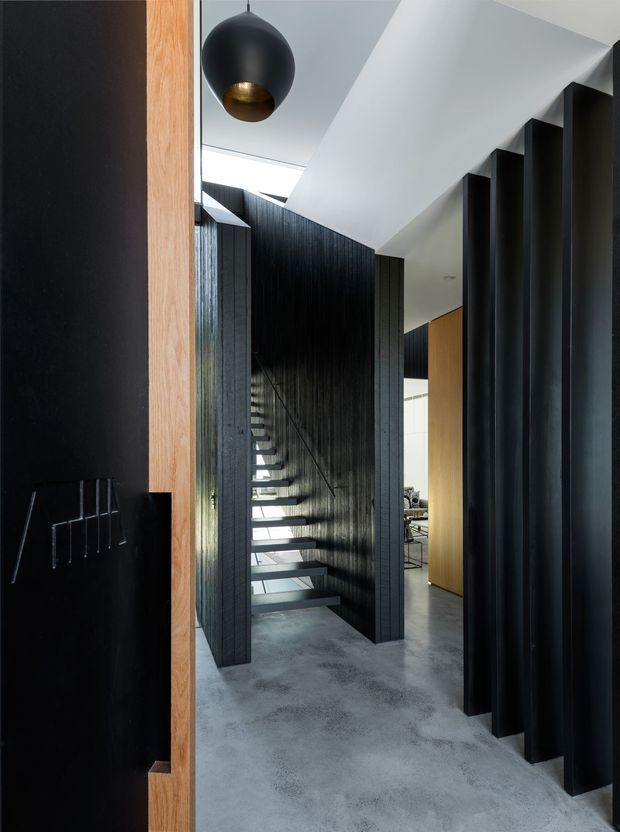 A dramatic contemporary interior hallway centred on a floating staircase. Black vertically ribbed wall panels and deep black structural fins contrast with a pale sculptural ceiling, polished grey concrete floor, and warm timber door framing. A black-and-brass spherical pendant hangs near the entry, while a glimpse of a furnished room is visible beyond the corridor.