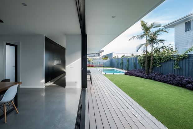 Contemporary indoor-outdoor living area opening onto a long timber-look deck and landscaped backyard. The interior has a polished grey concrete floor, white walls and ceiling, dark built-in cabinetry, and a small dining setting. Full-width retractable glazing creates a seamless connection to the deck, lawn, blue boundary fencing, planting and a compact swimming pool. A neighbouring weatherboard house is visible beyond the garden.