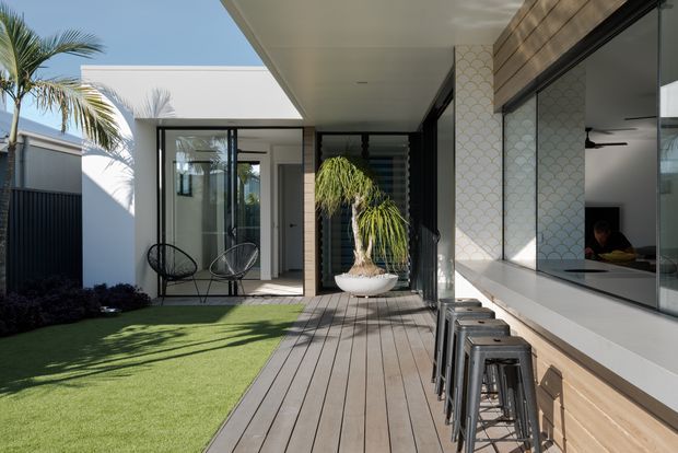 A contemporary outdoor entertaining area beside a modern home, featuring a timber-look deck, built-in outdoor bar with stools, large black-framed glazing, patterned tiled columns, artificial lawn, tropical planting, and a covered roof with a skylight or open section. The space is bright and relaxed, with seamless indoor-outdoor connection and a compact courtyard-style layout.