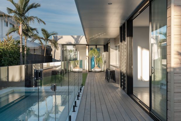A bright contemporary outdoor terrace or balcony alongside a narrow swimming pool, enclosed by frameless glass balustrades. The space features grey decking, extensive floor-to-ceiling glazing, dark metal framing, pale timber wall cladding, recessed ceiling lights, and tropical palm planting visible around the property.