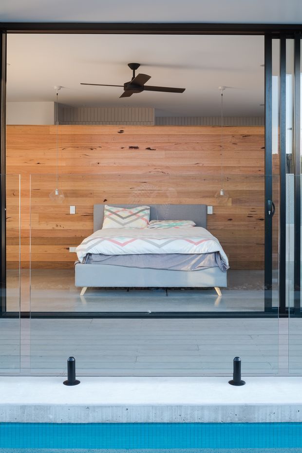 A modern bedroom viewed through a large glazed opening, with a low upholstered bed positioned against a full-height warm timber feature wall. The room has pale flooring, minimalist pendant lights, a dark ceiling fan, and dark-framed glazing. A swimming pool and concrete pool edge are visible in the foreground outside.