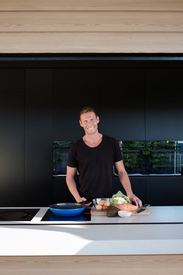 A contemporary kitchen interior photographed from across a large island. A person stands behind the island preparing food, with dark matte built-in cabinetry, a long horizontal window or glazed backsplash, a white island benchtop with an integrated black cooktop, and light timber wall and island cladding creating strong warm-and-dark contrast.