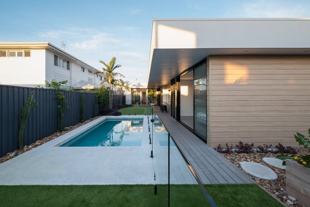 A contemporary single-storey residence viewed from its landscaped rear pool area. The image shows a long narrow swimming pool, pale concrete paving, dark metal boundary fencing, artificial lawn, a timber-look deck and walkway, extensive glazing, and a projecting flat-roofed white volume with horizontal timber cladding. Palm trees and neighbouring homes are visible in the background.