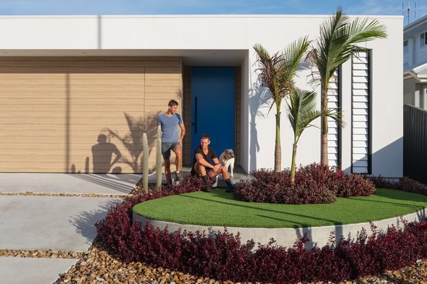 A contemporary single-storey house facade photographed from the front, featuring a broad white rendered wall, a large horizontal timber-clad garage door, a vivid blue entry door, and tall narrow window openings with horizontal screens. A curved raised garden bed with artificial-looking green lawn and burgundy planting defines the landscaped frontage, while two people and a dog stand near the entrance.