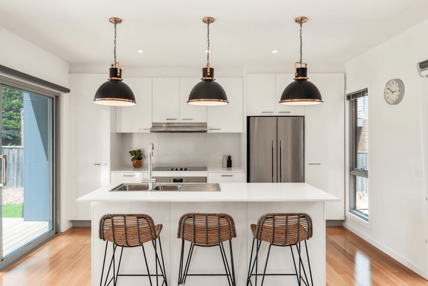 Bright contemporary kitchen with a large white island, three woven bar stools, white flat-panel cabinetry, stainless-steel appliances, and three black pendant lights. Timber flooring, extensive glazing, and a sliding glass door create an airy, clean atmosphere.