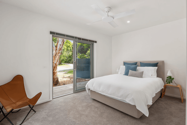 Bright, contemporary bedroom with a low upholstered bed dressed in white bedding and layered blue-grey cushions. The room has white walls, soft grey carpet, a ceiling fan, recessed lighting, and a large sliding glass door opening to a leafy outdoor area. A tan leather sling chair and compact bedside table add warm accents.