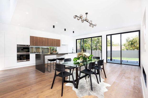 Bright open-plan kitchen and dining area with white floor-to-ceiling cabinetry, a dark stone island, warm timber upper cabinetry, timber flooring, and a black dining table with chairs. Large black-framed glazed doors open to an outdoor area, while a sculptural pendant light hangs above the dining zone. The space has a clean, contemporary appearance with abundant natural light.