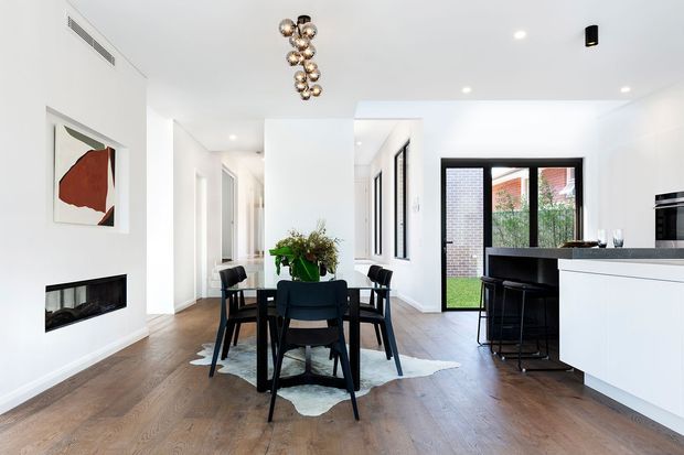 Bright open-plan contemporary dining and kitchen interior with white walls, warm timber flooring, a black-framed glazed door, and minimalist white cabinetry. A dark dining table with upholstered chairs sits on a pale hide rug, while a black kitchen island and bar stools occupy the right side. A linear fireplace, abstract artwork, indoor greenery, and a sculptural clustered pendant light add focal details.