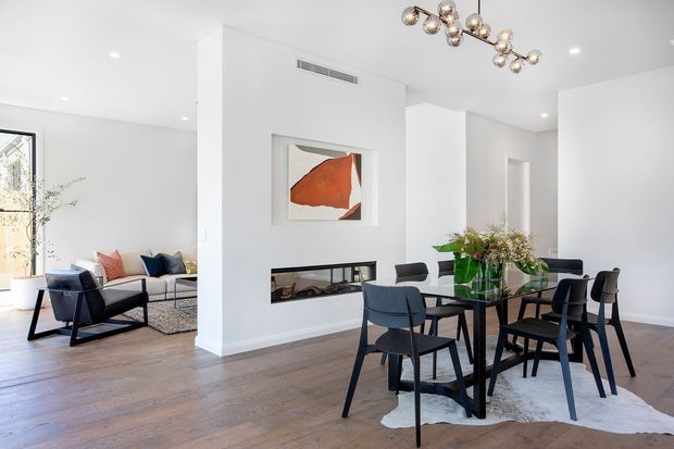 Bright open-plan contemporary interior combining a dining area and living room. The space features white walls and ceiling, wide warm-toned timber flooring, a built-in linear fireplace with dark surround, black-framed dining furniture, a sculptural globe chandelier, and an abstract artwork above the fireplace. A lounge seating area is visible to the left, with abundant natural light and restrained modern furnishings.