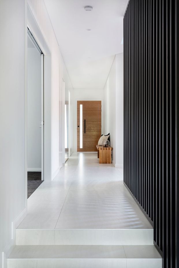 A bright, minimalist residential hallway and entryway with white walls, ceilings, and large-format pale flooring. Two shallow steps lead up to a corridor terminating in a warm timber pivot door with a narrow vertical glazed inset. A dramatic full-height black vertical slatted screen lines the right side, while white doorways and a glimpse of carpeted room appear on the left. A small timber bench with cushions sits near the entry door.