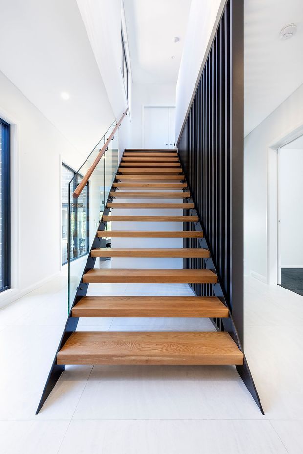 Bright contemporary stairway with open-riser timber treads supported by a dark metal structure. A clear glass balustrade with timber handrail runs along one side, while a tall black vertical-slat screen forms the other side. White walls, ceiling and large light floor tiles create a clean, airy interior; an adjacent room is visible through the doorway on the right.