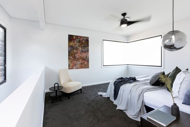 Bright contemporary bedroom with white walls and ceiling, dark charcoal carpet, a large bed dressed in white and grey bedding, and a sculptural upholstered armchair beside a small black side table. A corner window provides strong natural light, while a polished spherical pendant and ceiling fan add modern features. Abstract artwork and restrained furnishings create a clean, refined atmosphere.